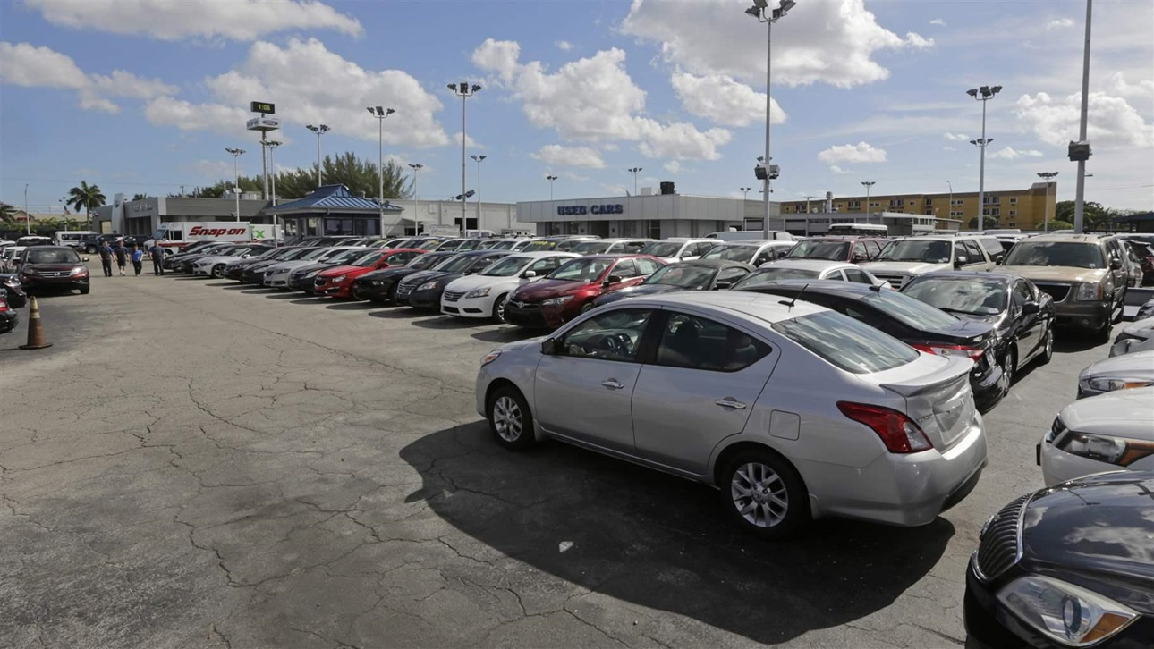 A used car dealership. Many modern cars are lined up in neat rows. The asphalt of the lot has cracks in it.