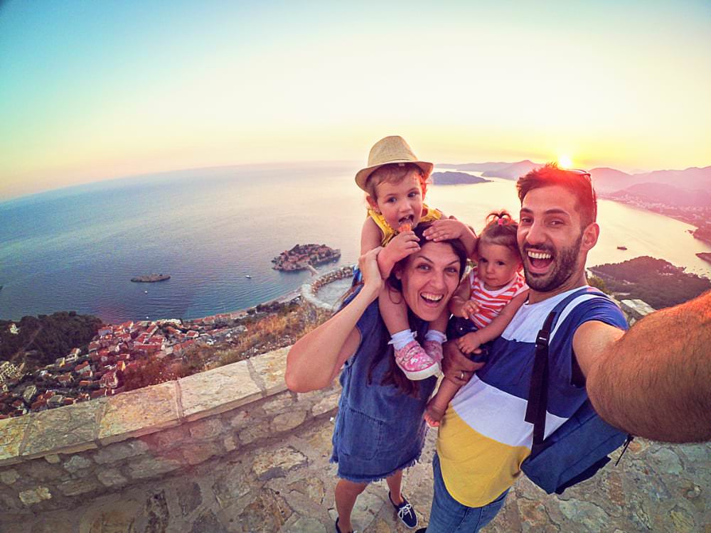 A family of four smiling while standing on top of a scenic vacation outlook. The father is taking a selfie while holding his daughter. The mother has her son on her back, and is standing next to him. Behind them the sun is gently setting over an ocean villa.